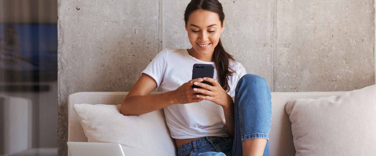 Happy young asian woman using mobile phone while sitting a couch at home with laptop computer