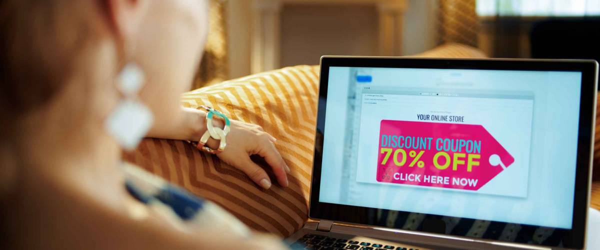 Over shoulder shot of modern woman reading e-commerce email with discount coupon on a laptop laying on sofa at modern home in sunnny hot summer day.