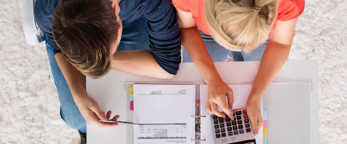Couple Using Calculator For Calculating Invoice, Tax, Bills And Retirement Money On Desk At Home