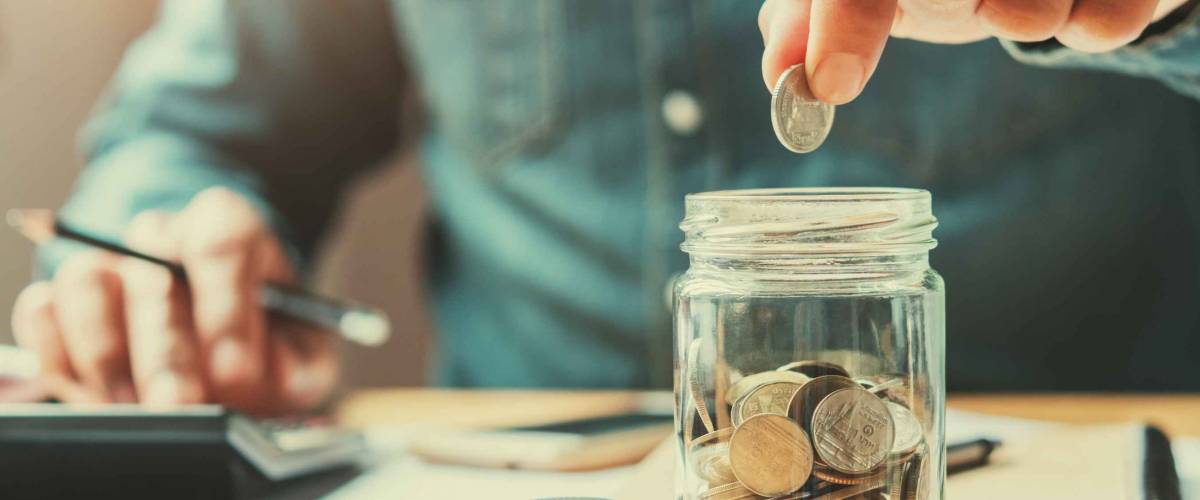 man drops coins into glass jar, with other hand on a calculator