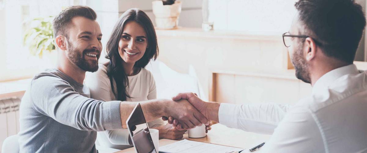Good deal! Cheerful young man bonding to his wife while shaking hand to man sitting in front of him at the desk