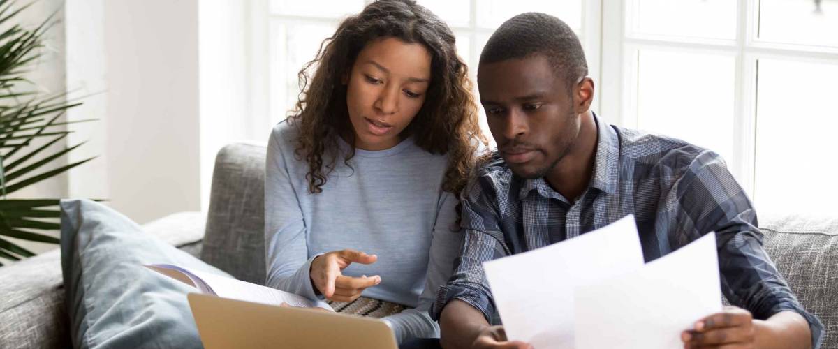 Serious African American couple discussing paper documents, sitting together on couch at home, man and woman checking bills, bank account balance, terms of contract, mortgage, loan agreement