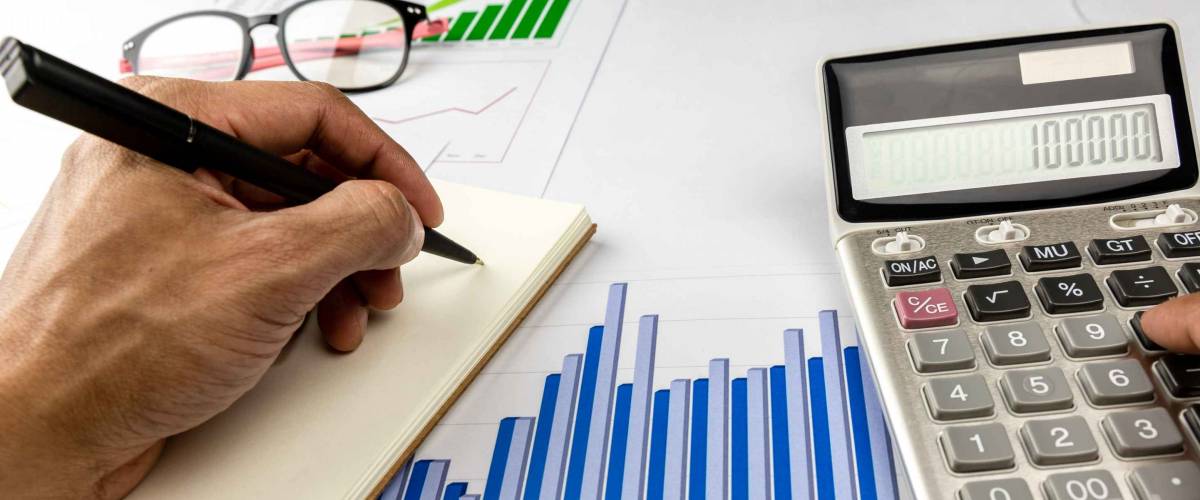 close up of calculator, graphs, glasses on a desk, while a man jots something in a notebook