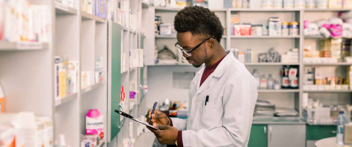 Young African American man pharmacist standing in interior of pharmacy. Man specialist of pharmacy making notes on clipboard during inventory