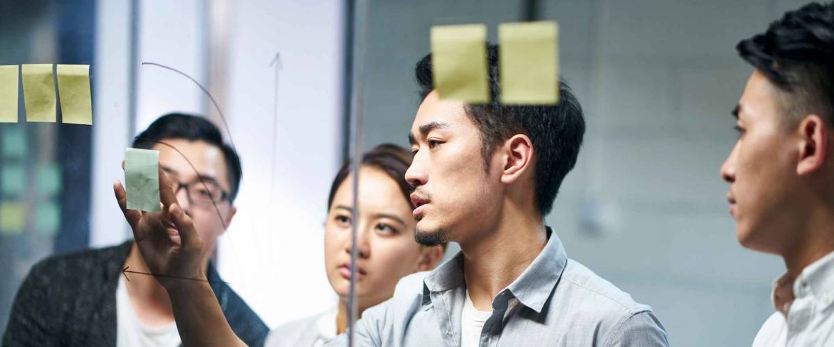 young asian entrepreneur of small company putting a adhesive note on glass in office during team meeting formulating business strategies.