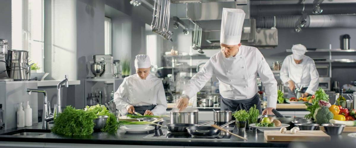 Famous Chef Works in a Big Restaurant Kitchen with His Help. Kitchen is Full of Food, Vegetables and Boiling Dishes.