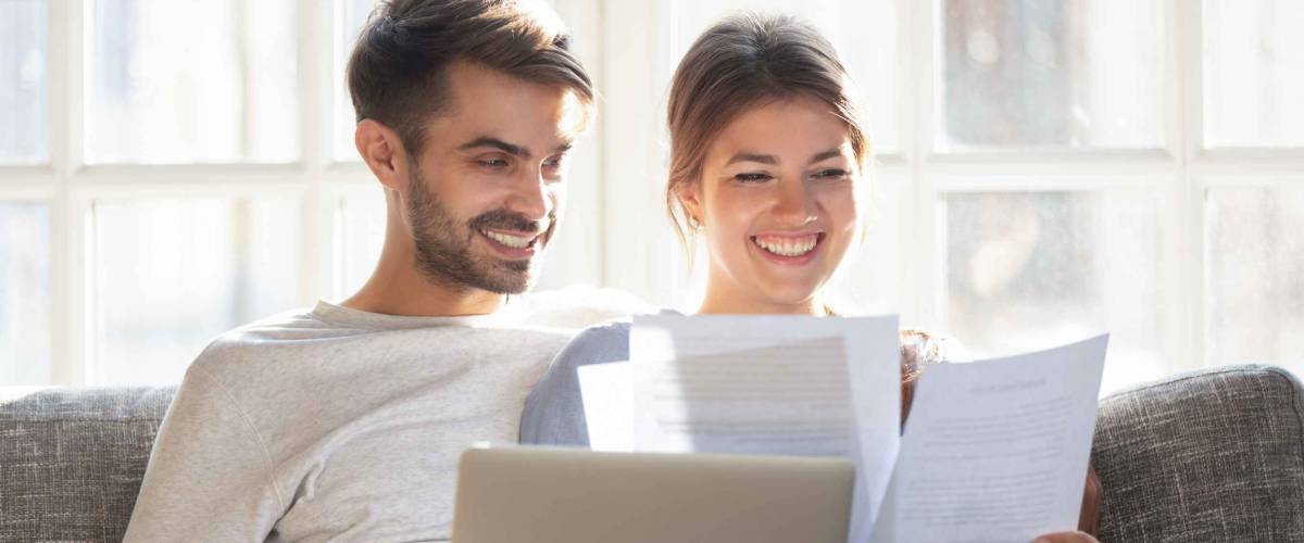 smiling man and woman sit on a couch and compare papers