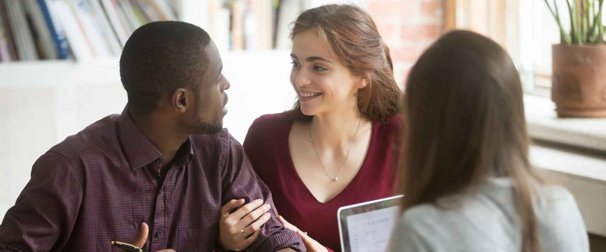 young couple sit at desk across from woman and talk to each other, making a decision
