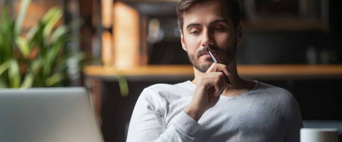 Pensive bearded man sitting at table drink coffee work at laptop thinking of problem solution, thoughtful male employee pondering considering idea looking at computer screen making decision