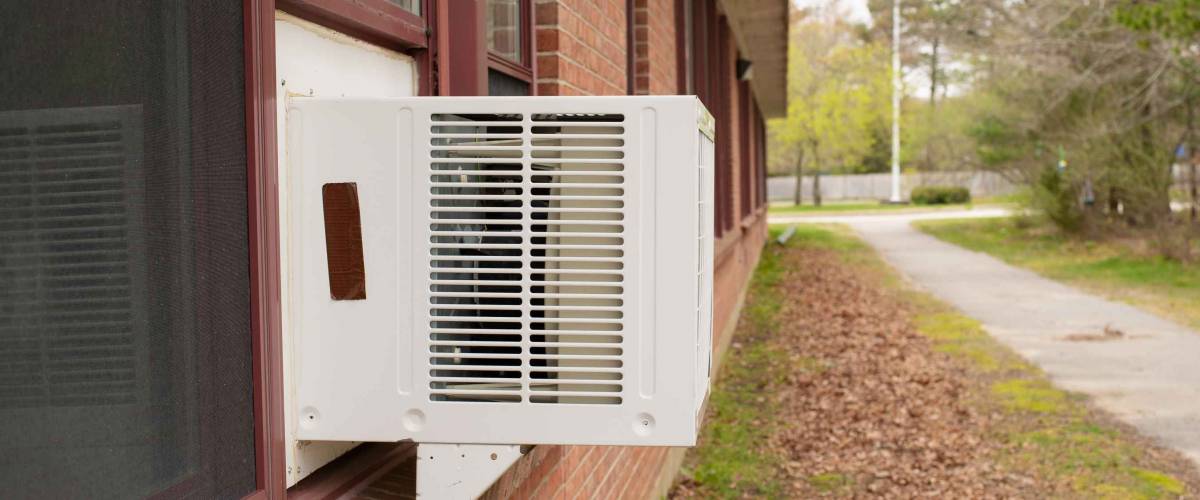 Window air conditioner unit in window in brick building with green grass and leaves on ground on overcast day
