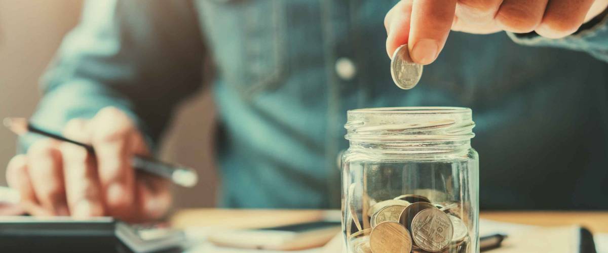 man puts coin in glass jar, while using calculator with other hand