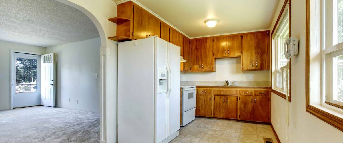 empty home, older cabinets in the kitchen, beige carpeting in the living room adjacent