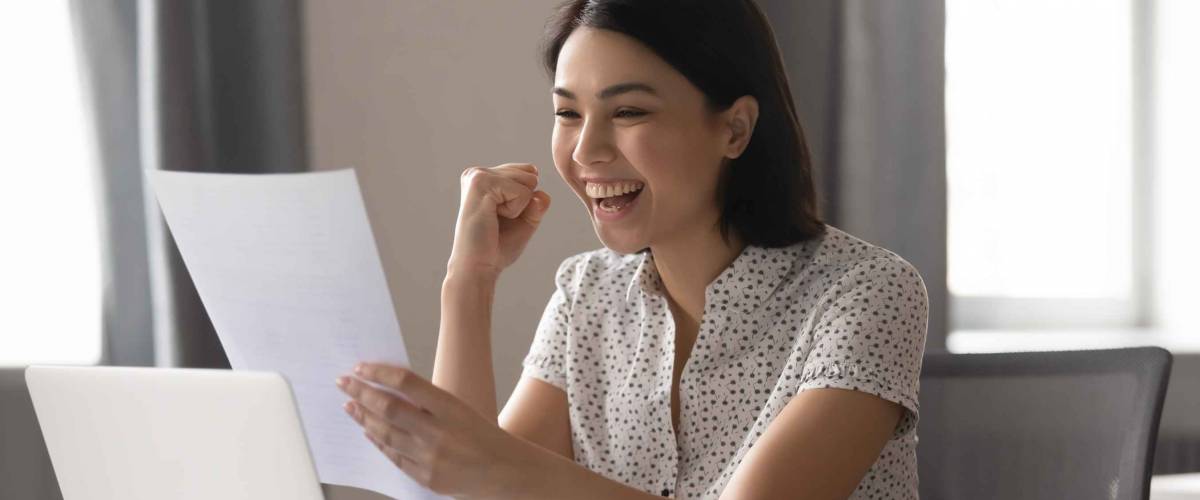 Happy woman looks at sheet of paper in front of laptop