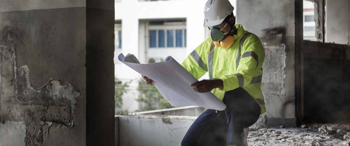 man in hardhat and safety gear looks at plans on construction site