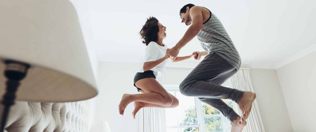 Man and woman holding hands and jumping together on bed. Couple having fun in bedroom.