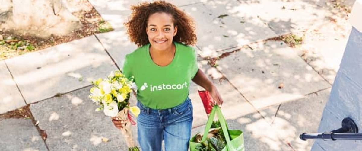 woman wearing instacart tshirt walks with bag of groceries and