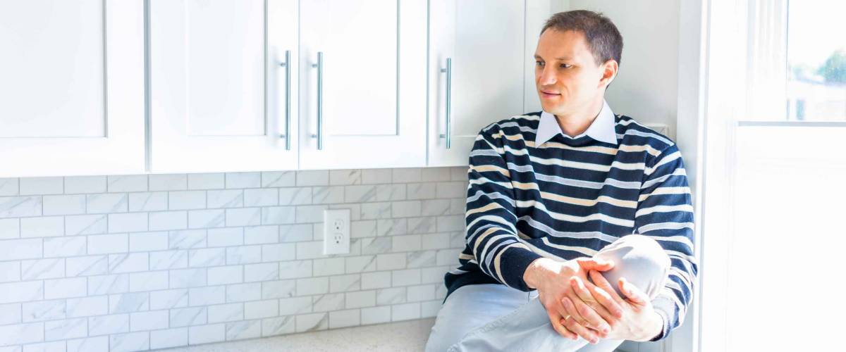 Young happy man sitting on modern new kitchen countertop by window smiling
