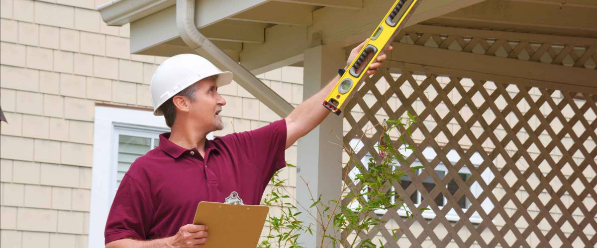A home inspector or house building repair contractor in a hard hat holding a level and a clipboard outside a home doing an inspection or construction quote