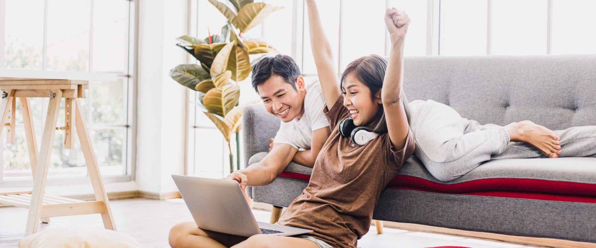 Happy Asian couple shopping online with laptop computer at home. Lovely couple making purchases online or booking in beautiful living room. Buying ordering products on the internet.