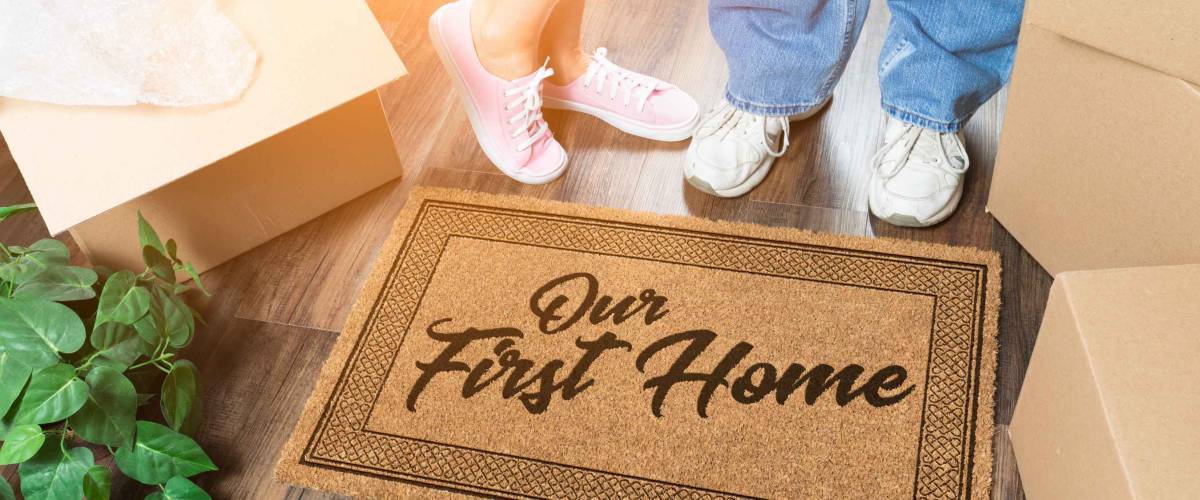 Man and Woman Unpacking Near Our First Home Welcome Mat, Moving Boxes and Plant.