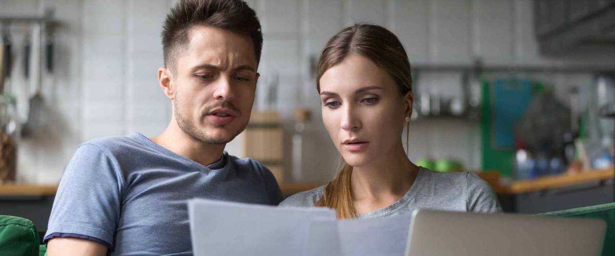 Serious looking man and woman sitting on couch, looking at papers and laptop