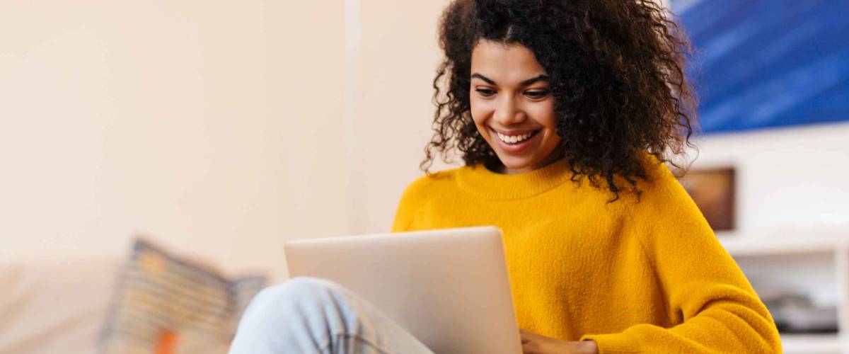Image of cheerful african american woman using laptop while sitting on chair in living room