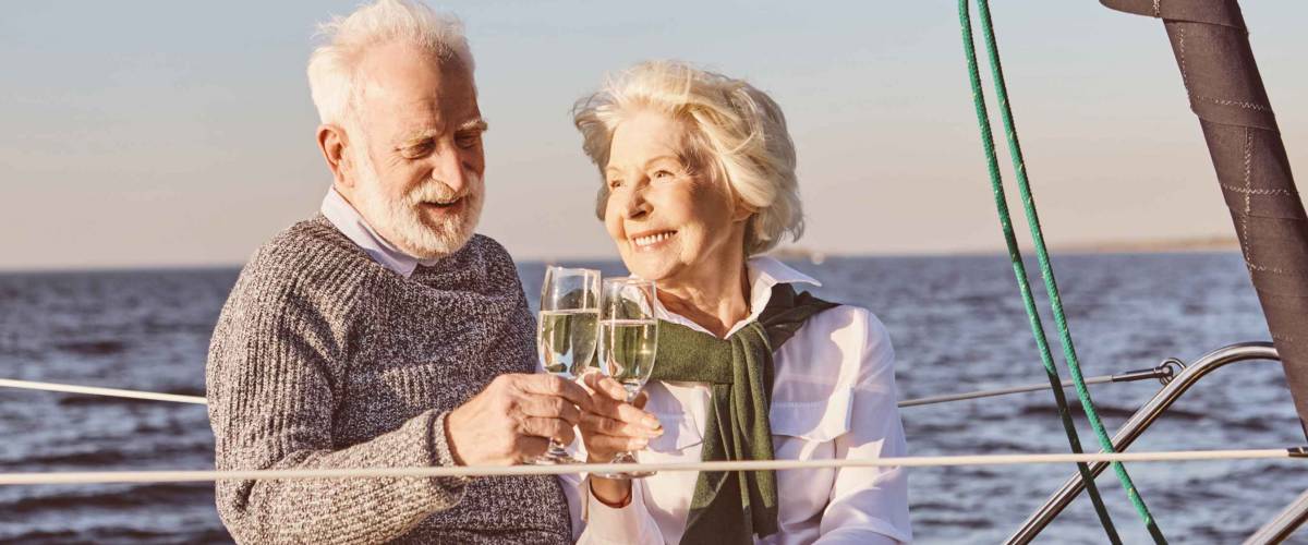 Cheers. Close up of happy senior couple sitting on the side of sail boat or yacht deck floating in sea. Man and woman drinking wine or champagne, enjoying the view