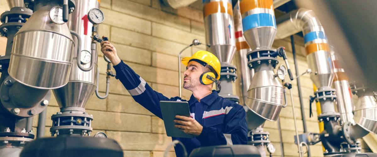 Young Caucasian worker in protective suit tightening the valve and using tablet while standing in heating plant.