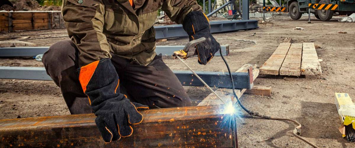 A young man welder in brown uniform, welding mask and welders leathers, weld metal with a arc welding machine at the construction site, blue sparks fly to the sides