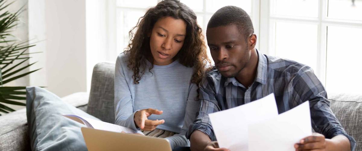 couple discussing paper documents, sitting together on couch at home, in front of computer