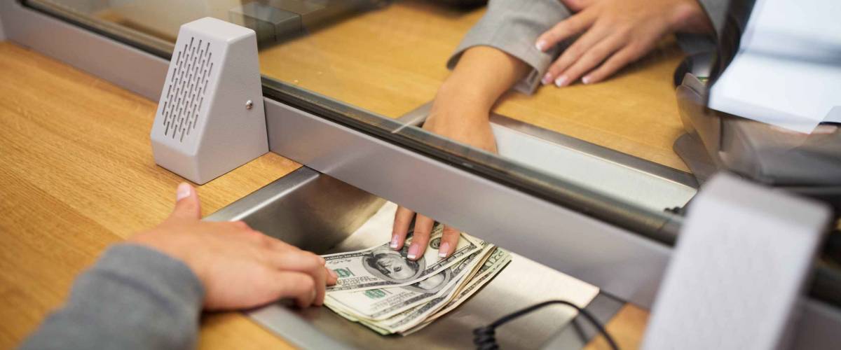 person taking cash from teller at bank under glass partition