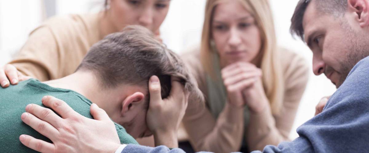 Close-up of young man holding his head in his hands and a group of friends in a supportive pose around him