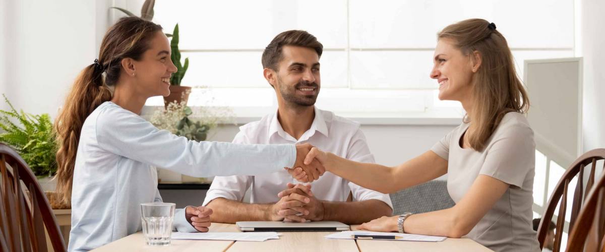 Two women shake hands across a table, smiling, while man sits in between, also smiling
