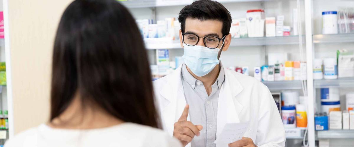 Middle eastern male pharmacist wearing protective hygienic mask to prevent infection selling medications to woman patient to prescription and making drug recommendations in modern pharmacy