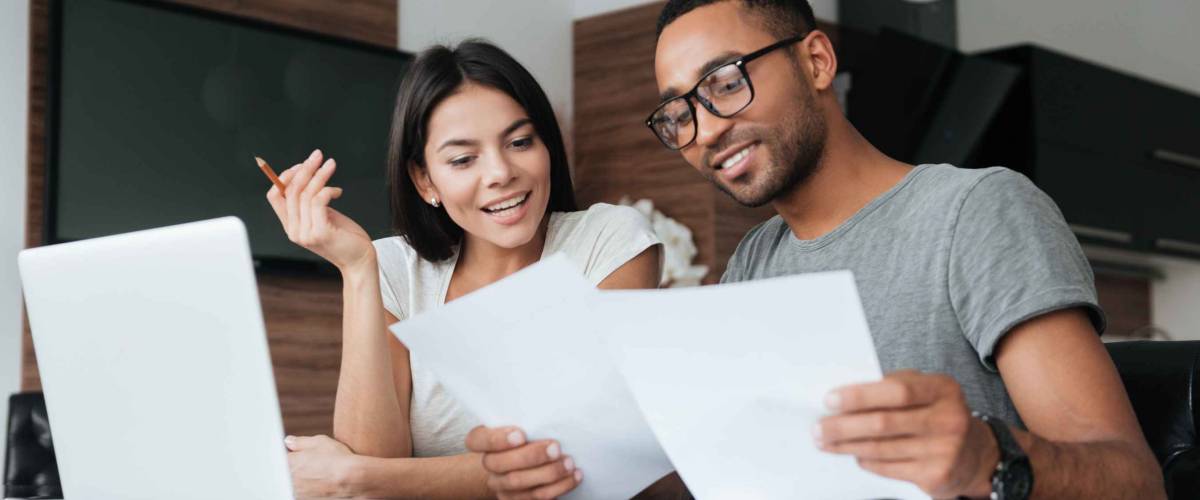 Photo of cheerful loving young couple using laptop and analyzing their finances with documents. Look at papers.