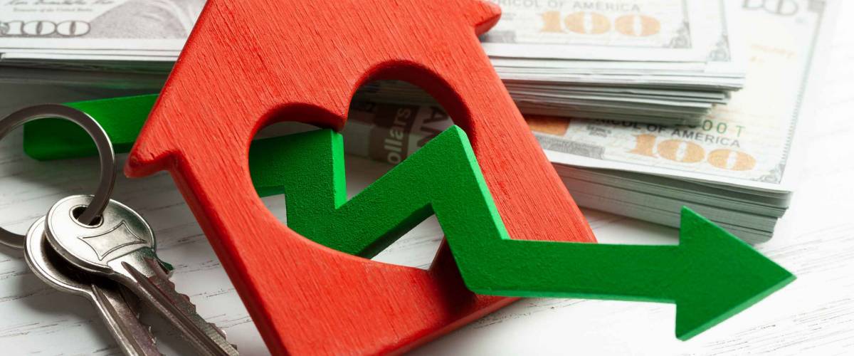 Arrow down and house with money on white wooden background. Falling market price of real estate.