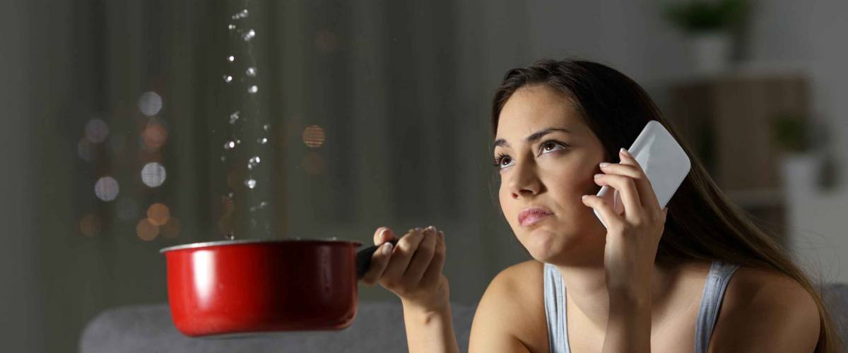 Frustrated young woman holding out a pot to catch falling water droplets, holding phone up to her ear
