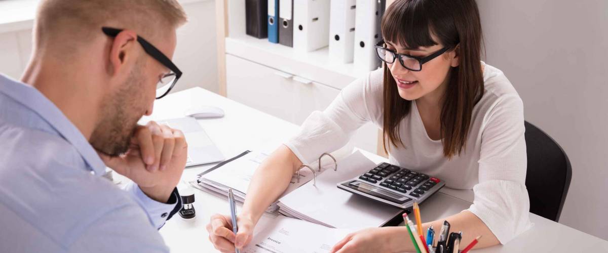 Woman sitting at desk shows man across from her something on a piece of paper