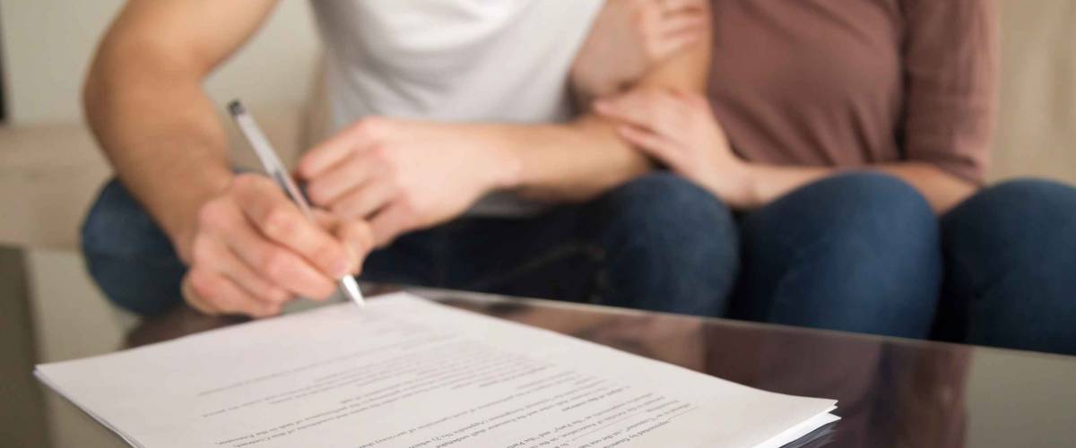 Close up of signing loan agreement, couple sitting on sofa, male hand with a pen setting signature, taking bank credit with easy terms of payment and low interest rate for purchasing property