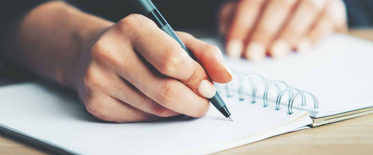 Close up of woman's hands writing in spiral notepad placed on wooden desktop with various items