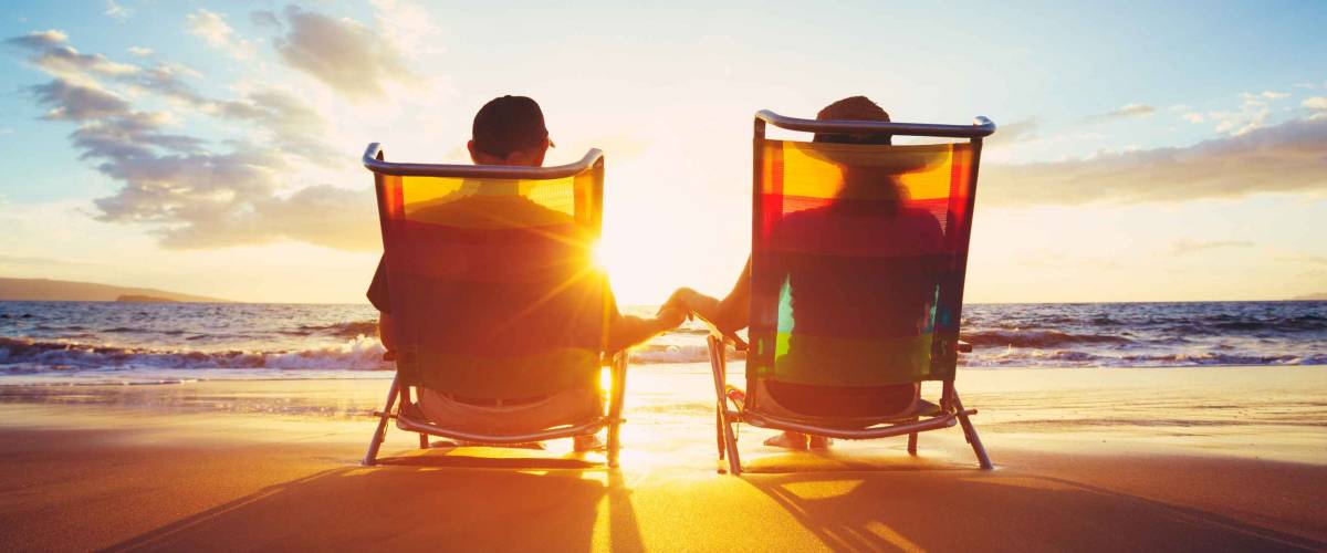 Older couple sitting on a beach in chairs, seen from behind as the sun sets