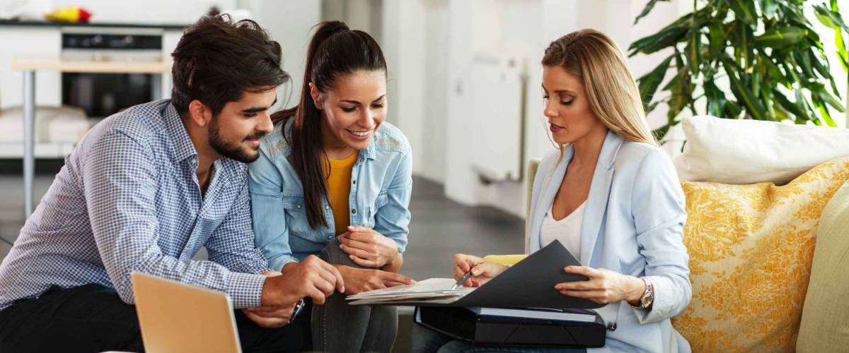 Woman in suit shows young couple something in binder, sitting in a house together