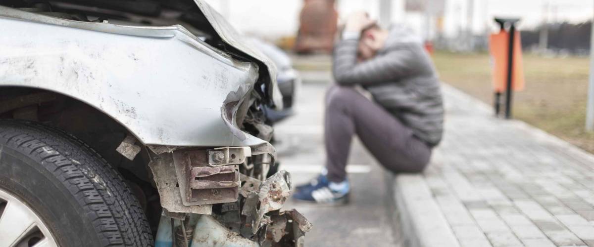 broken car after the accident in foreground, upset man sitting on curb