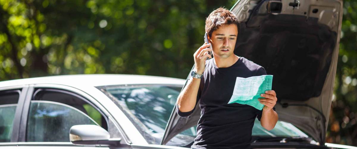 Young man leaning against car with open hood, on the phone and looking at a sheet of paper