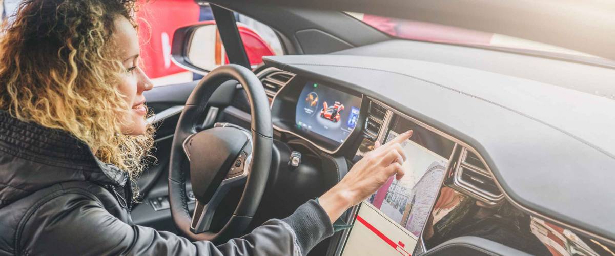 Young woman sits behind wheel in car and uses an electronic dashboard.