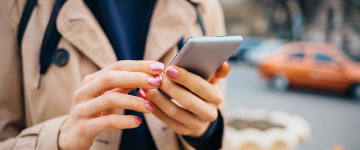 Young woman wearing beige coat using smart phone standing near road with cars in city at spring day. Close-up female's hands holding mobile phone outdoors.