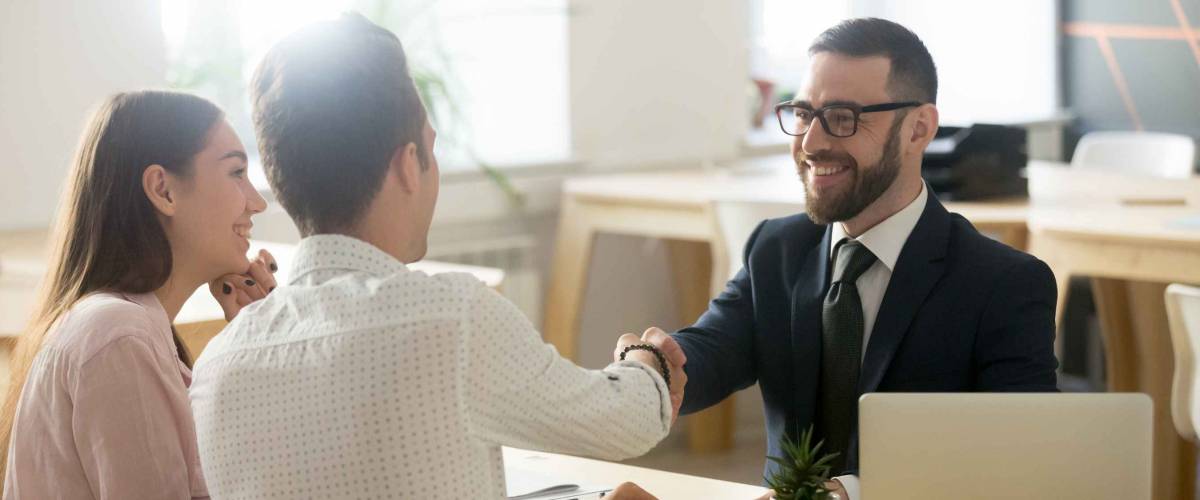 Smiling man in suit shakes hands with young couple sitting across the table from him