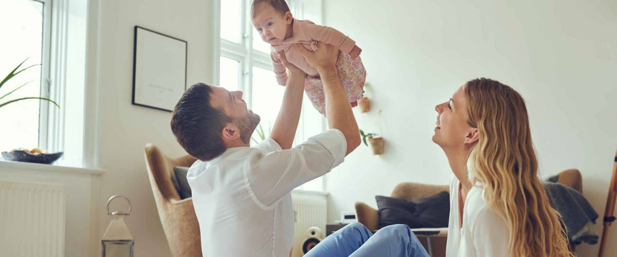 Proud father holding his newborn baby daughter in the air, with smiling mother sitting on the floor at home