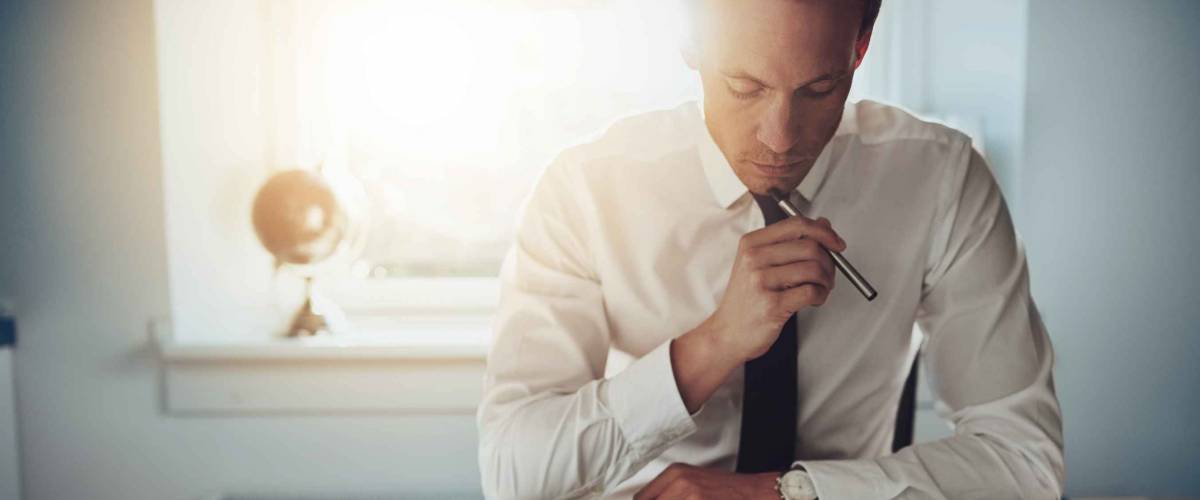 Serious looking man in shirt and tie sits at desk and looks down with pen held to his chin