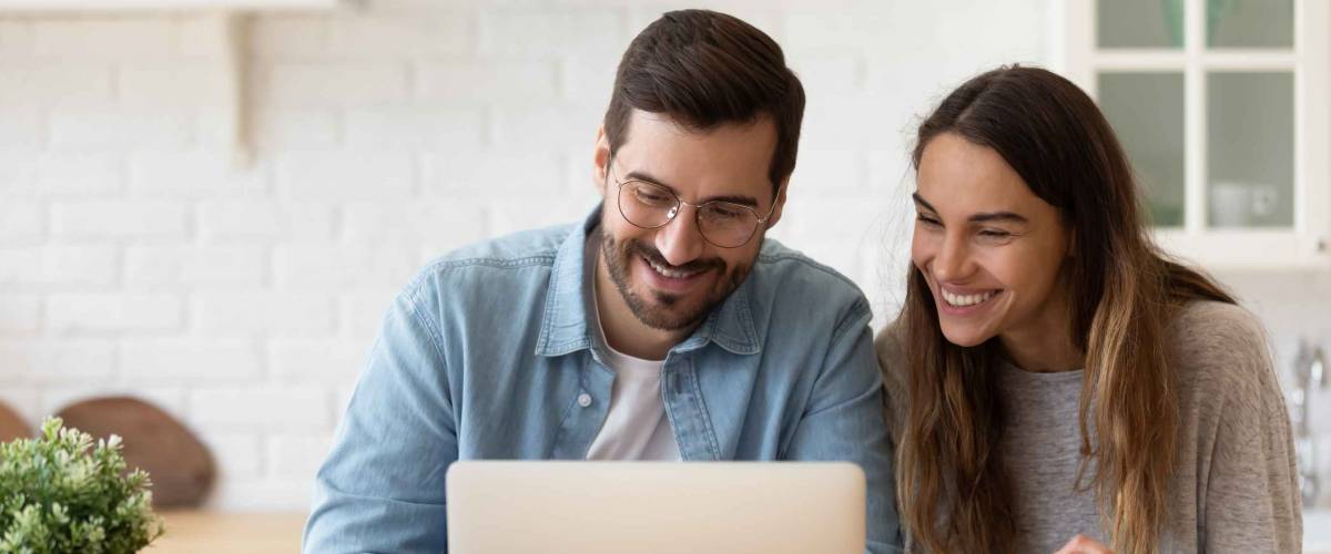 Young couple sitting at kitchen table, smiling, looking at laptop